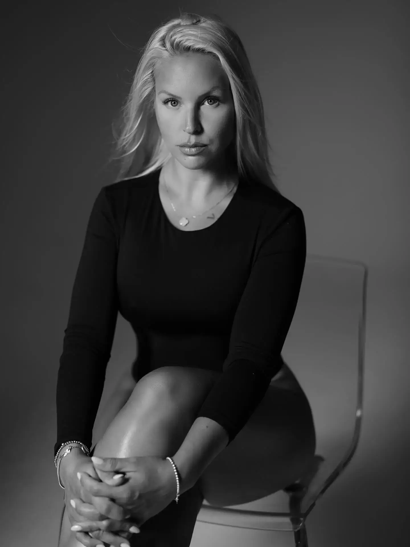Chosen By Ekaterina Black and white photo of a woman with long blonde hair, wearing a long-sleeve top, sitting on a transparent chair with her hands and legs crossed, looking at the camera—capturing the essence of female strategy growth and development. Women & Female Mentorship